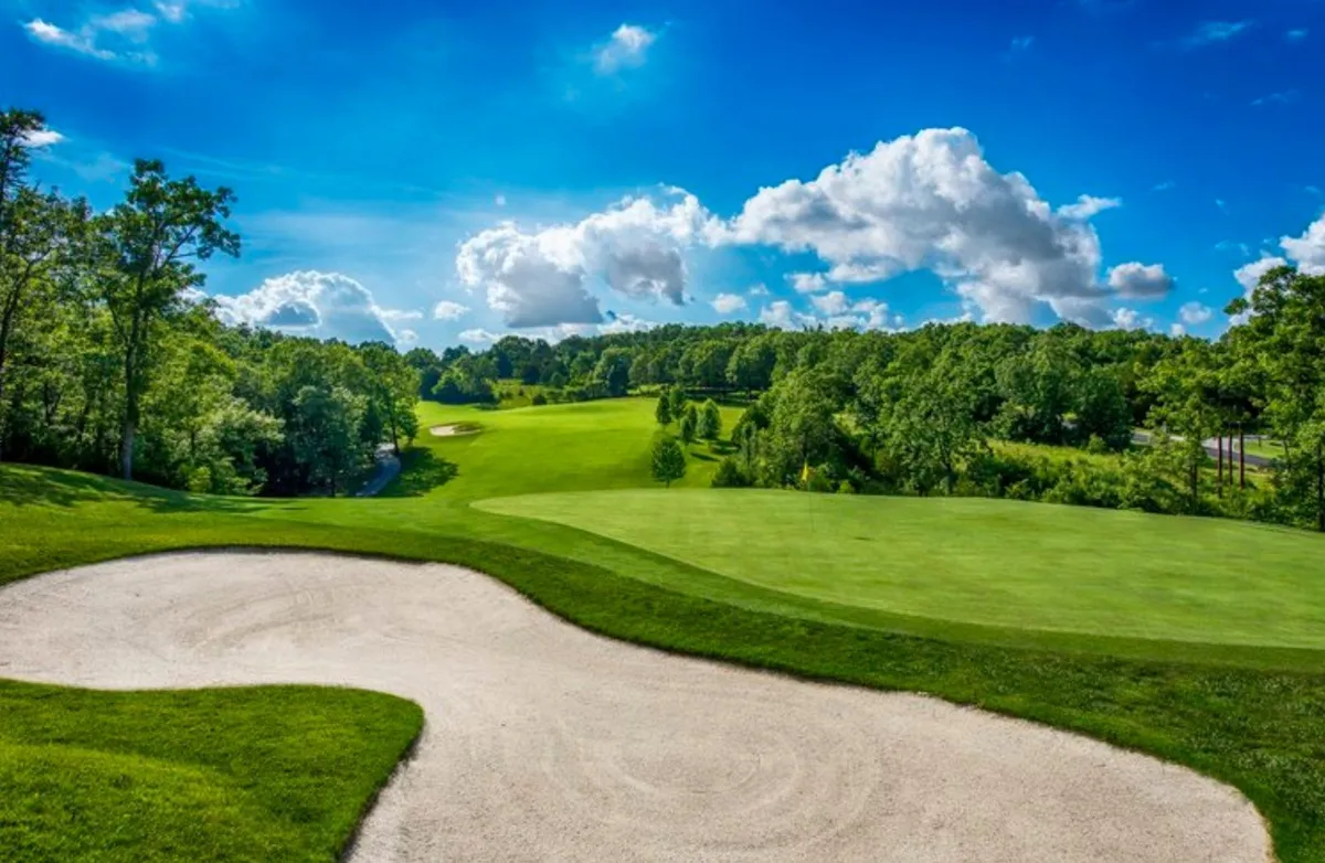 A lush green fairway with a sand bunker beneath a wide blue sky dotted with clouds. The expansive course is inviting and reflects the 'masterpiece of mountain golf architecture' referenced, set against a scenic natural backdrop that evokes the Ozark setting.