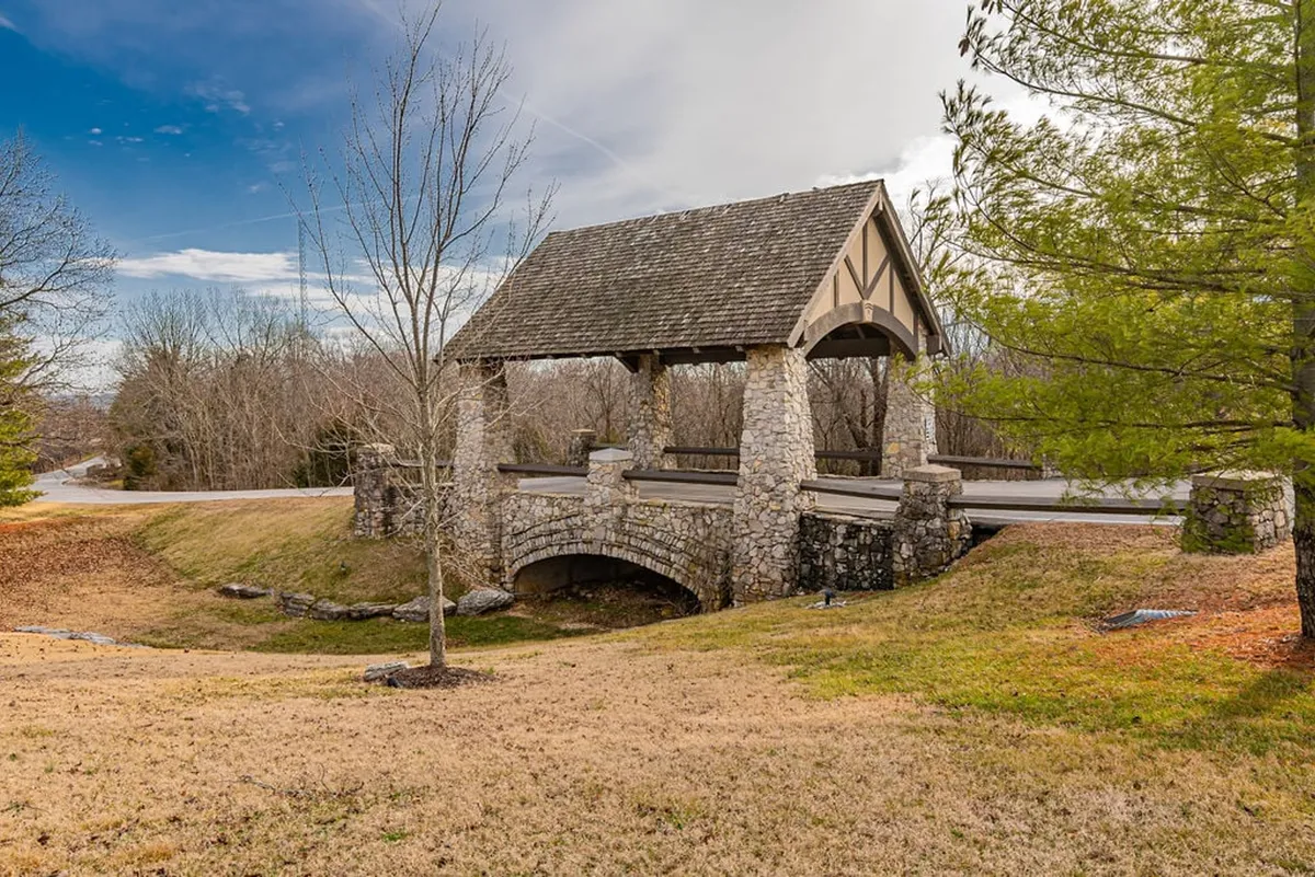 This peaceful exterior image features a charming stone bridge, a wooden shelter, and surrounding trees with an open grassy area. The mood is intimate and tranquil, inviting quiet walks and togetherness in nature.
