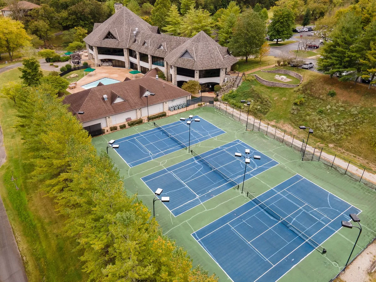 This exterior view showcases the tennis courts bordered by mature trees, with the resort clubhouse in the background. The scene is lively and social, hinting at friendly matches and summer events.