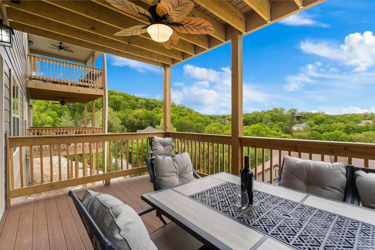 An exterior deck scene featuring a dining table with cushioned chairs, a ceiling fan, and a clear view of rolling green hills beyond the railing—ideal for both casual lounging and group meals.