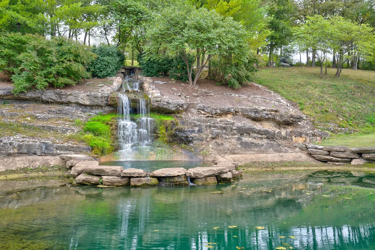 A tranquil natural pond with a small waterfall, surrounded by rock formations and lush greenery. The water is reflective, and the area appears peaceful and undisturbed—ideal for relaxation, fishing, or nature walks.