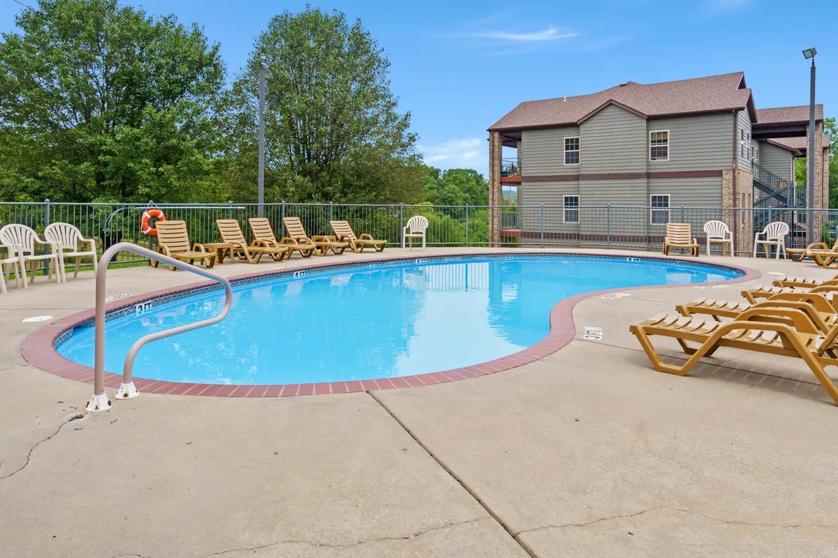 A sparkling outdoor swimming pool surrounded by lounge chairs and lush trees, with a building in the background. The pool area is bright and inviting, perfect for family relaxation and play during the warm months.
