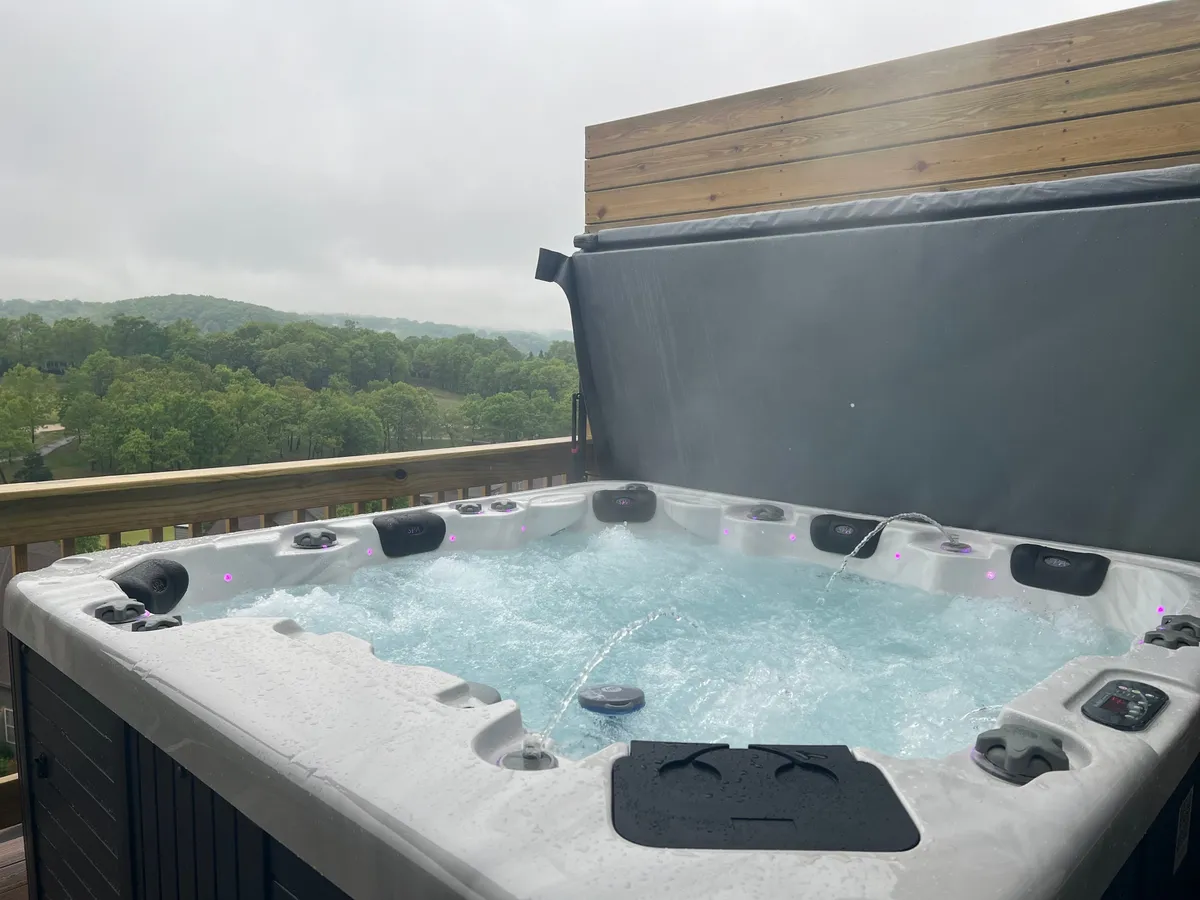 The hero image showcases the condo's private deck featuring a bubbling hot tub, set against a scenic backdrop of lush greenery and expansive views. The inviting hot tub is the centerpiece, with a sturdy deck railing framing the peaceful, natural surroundings. The mood is tranquil, evoking relaxation and the simple pleasures of unwinding outdoors at sunset or beneath the stars.