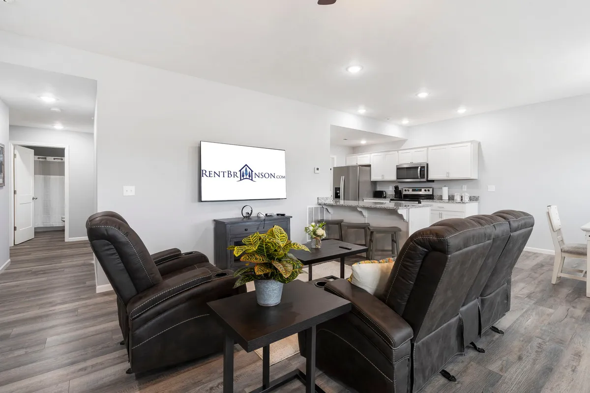 This image captures an inviting, open-concept living space with comfortable recliner chairs, a large wall-mounted TV, a kitchen area featuring stainless steel appliances, and a dining table. Natural light fills the room, and the layout encourages connection and relaxation.