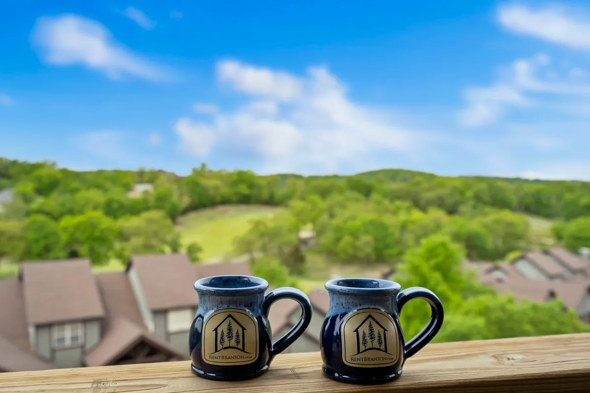 A serene balcony scene featuring two coffee mugs set on a table, with a lush green landscape and a clear blue sky in the background. The vantage point from the deck provides a calm, inviting place to relax and take in the peaceful views after a day of exploration.