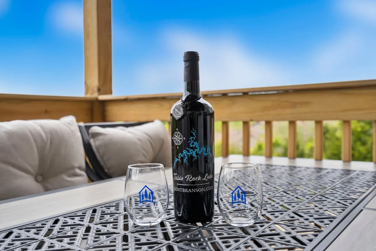 A cozy outdoor seating area with a bottle of wine and two glasses arranged on the table, set against a backdrop of open sky and landscape. The scene suggests a relaxed, intimate atmosphere perfect for enjoying a drink while taking in the sunset.