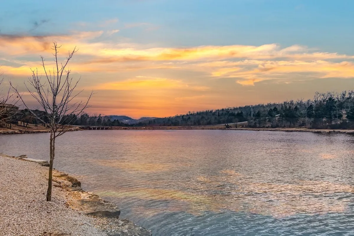 A picturesque lake scene at sunset, with the vibrant sky reflecting over calm waters. A tree frames the foreground, suggesting a peaceful moment perfect for fishing or simply enjoying the view.