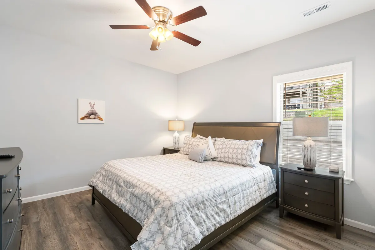 A serene king bedroom featuring a large bed with plush bedding, a nightstand with a lamp, tasteful artwork, and a window letting in soft sunlight. The setup invites relaxation and showcases a private, comfortable retreat ideal for couples or parents.
