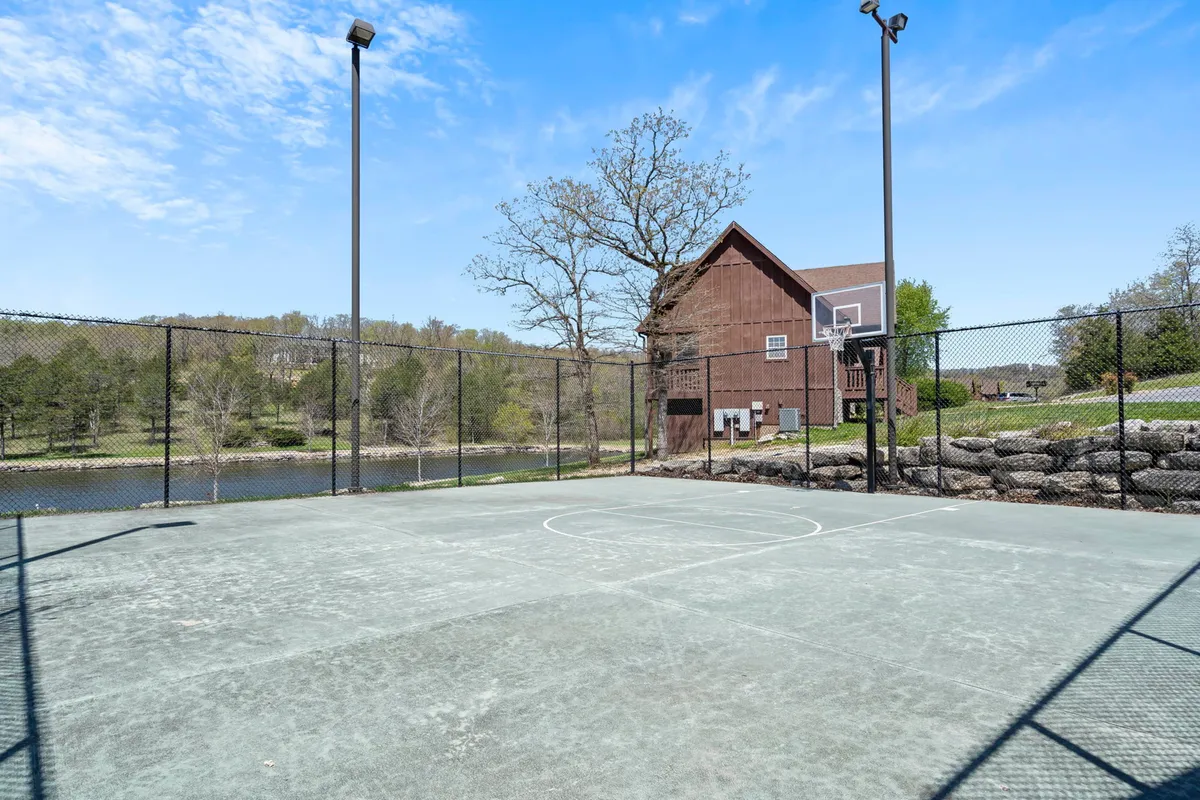An energetic basketball court set near a tranquil water body, surrounded by mature trees and a house in the background. The court is ready for games, while the setting invites both activity and relaxation.