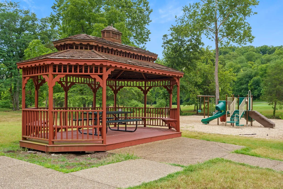 A cheerful playground set amidst green trees, featuring a gazebo and vibrant play equipment. Children can be seen enjoying the space while families gather for picnics or casual outdoor fun.